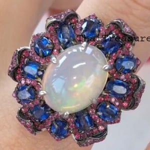 May include: A large, ornate ring featuring a central oval opal stone surrounded by blue sapphires and smaller pink gemstones. The ring has a floral design and is set on a silver band. The text "Thai Treasure" is visible on the ring.