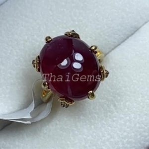 May include: A gold ring featuring a large, oval, deep red gemstone. The gemstone is set in a gold band with decorative detailing. The ring is presented in a red and white jewellery box. The text "Thai Gems" is visible on the gemstone.