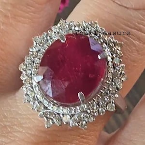 Op de afbeelding: Een opvallende ring met een grote, ovaalvormige robijn omringd door een halo van kleine diamanten. De robijn heeft een dieprode kleur en is gezet in een zilveren of witgouden band. Een opvallend sieraad.