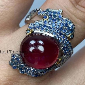Könnte beinhalten: Ein silberner Ring mit einem großen, runden, tiefroten Edelstein. Der Ring ist mit blauen Edelsteinen und kleinen goldenen Akzenten verziert. Der Text "ThaiTreas" ist auf dem Ring zu sehen.