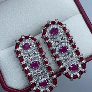 Puede incluir: Un par de pendientes rectangulares con borde rojo y tres gemas ovaladas de color rosa intenso. Los pendientes tienen un engaste plateado con pequeñas gemas transparentes alrededor de las piedras rosas. Los pendientes se muestran en una caja de joyería roja.