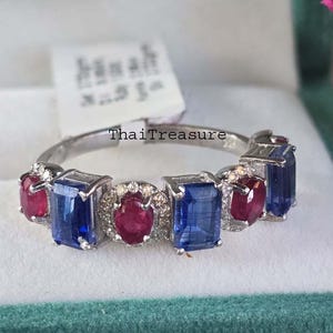 May include: A silver ring with alternating blue and red gemstones. The ring features rectangular blue stones and oval red stones, set in a band with the text "ThaiTreasure". The ring is displayed in a teal jewelry box.