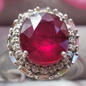 Puede incluir: Primer plano de un anillo de plata con una gema grande, redonda y de color rojo intenso, rodeada de piedras más pequeñas, transparentes y con corte baguette. El texto "ThaiTreasure" es visible.