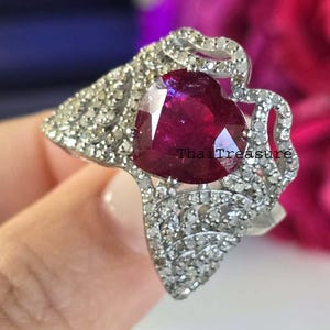 May include: A ring featuring a heart-shaped, deep red gemstone set in a silver-coloured band. The band is embellished with small, clear stones. The ring is held up by a hand. The text "Thai Treasure" is visible.