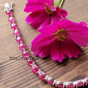 Pode incluir: Uma pulseira de rubi com pedras preciosas ovais e um fecho prateado é exibida sobre uma superfície de madeira, junto a duas vibrantes flores de cosmos cor-de-rosa. A pulseira é uma joia, e as flores adicionam um toque de beleza natural.