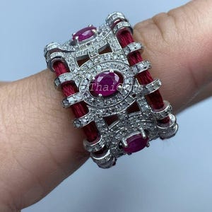 Peut inclure: Une bague en argent ornée de trois pierres précieuses ovales en rubis et de nombreux petits diamants. La bague présente un motif de cordon rouge et le texte "Thai Gems".