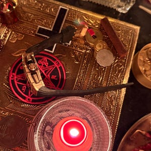 May include: A gold-toned wristwatch with a black rubber strap featuring a prominent crown and side detailing, resting on a rectangular gold-coloured metal plate engraved with a black cross and a red circular emblem with black symbols.