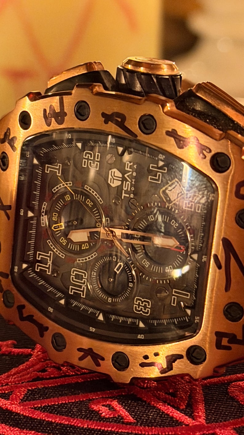 May include: A copper-toned tonneau-shaped wristwatch with a black dial featuring the text TSAR BOMBA. The bezel displays black engraved symbols, and the watch includes three sub-dials, luminous hands, and large Arabic numerals for the hours 1, 10, 11, and 12.