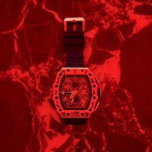 May include: A black wristwatch with a tonneau-shaped case featuring a bezel engraved with dark symbols. The watch has a black silicone strap and a complex dial with multiple sub-dials, all illuminated under a vibrant red light source.