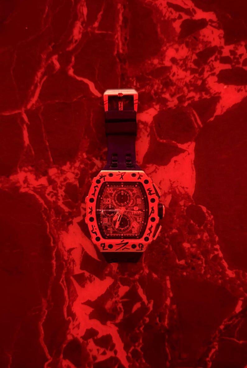 May include: A black wristwatch with a tonneau-shaped case featuring a bezel engraved with dark symbols. The watch has a black silicone strap and a complex dial with multiple sub-dials, all illuminated under a vibrant red light source.