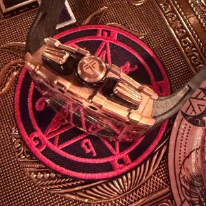 May include: A metallic watch with a copper-toned case and a black rubber strap, featuring a prominent crown with a circular logo. The watch rests on a circular patch with a red and black geometric design, surrounded by various metallic coins.