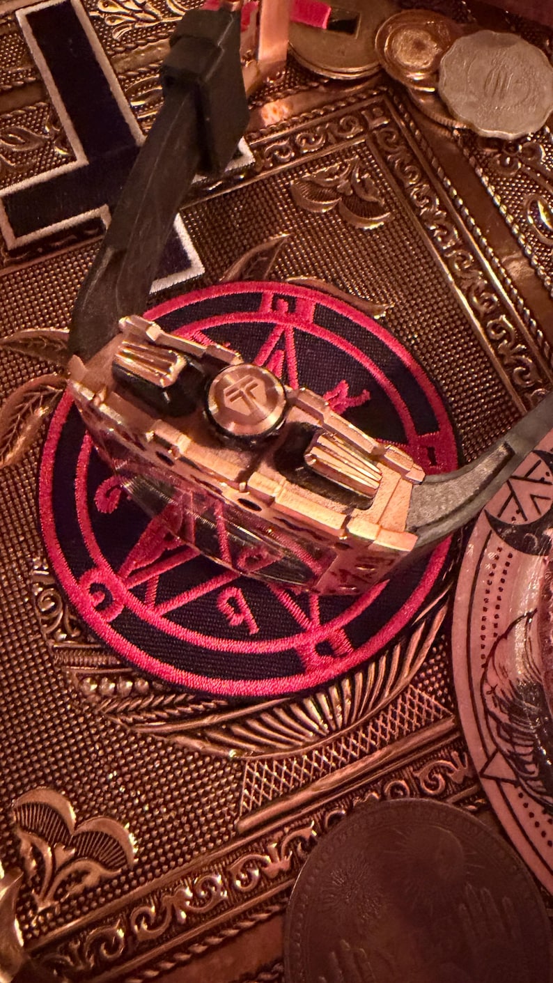 May include: A metallic watch with a copper-toned case and a black rubber strap, featuring a prominent crown with a circular logo. The watch rests on a circular patch with a red and black geometric design, surrounded by various metallic coins.