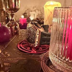 May include: A luxury wristwatch with a gold-toned tonneau-shaped case and a black rubber strap, resting on a circular black and red pentagram coaster. The watch features a complex black dial with multiple sub-dials and gold-toned accents.