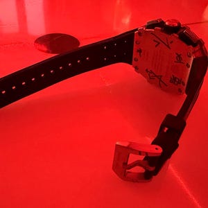 May include: A black wristwatch with a perforated silicone strap and a silver-toned metal buckle. The watch case back is silver-toned and features black engraved markings and symbols, with a prominent red circular detail visible on the casing.