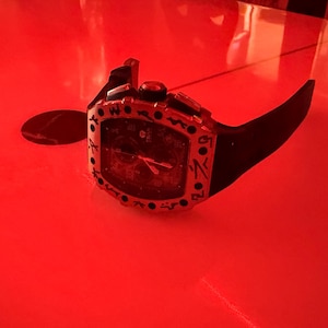 May include: A black wristwatch with a tonneau-shaped case featuring a silver-toned bezel with black engraved markings. The watch has a black dial with visible hands and a black rubber strap, presented under a strong red light source.