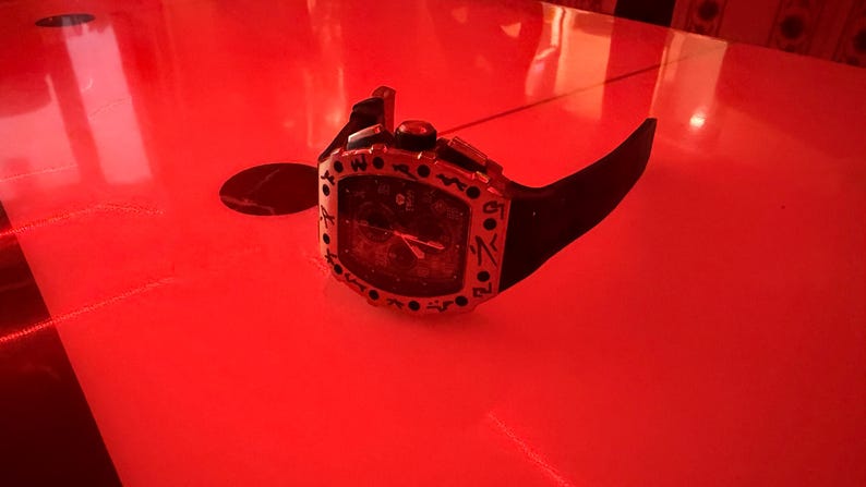 May include: A black wristwatch with a tonneau-shaped case featuring a silver-toned bezel with black engraved markings. The watch has a black dial with visible hands and a black rubber strap, presented under a strong red light source.