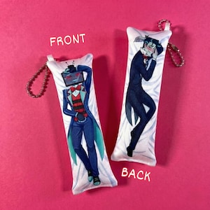 May include: Two white body pillow keychains with illustrations on a pink background. One keychain shows a character with a TV for a head, wearing a blue suit. The other keychain shows a character in a black suit. The word "FRONT" and "BACK" are printed on the pillows.