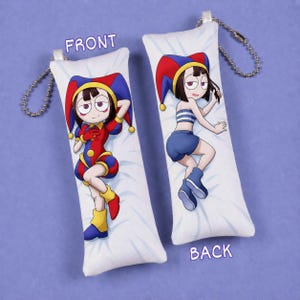 Puede incluir: Dos llaveros de peluche rectangulares con un personaje de dibujos animados de pelo oscuro y gorro de bufón. El llavero de la izquierda muestra al personaje con un traje de bufón rojo y azul. El de la derecha, con top de rayas azules y pantalones cortos.