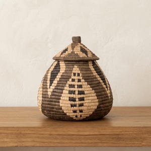 May include: A woven lidded basket with a rounded base and a pointed top. The basket features a repeating geometric pattern in dark brown and natural beige fibers, creating a series of leaf-like shapes around the body and the lid.