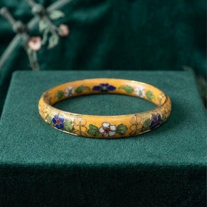 May include: A yellow enamel bangle bracelet featuring floral patterns in white, blue, and green. The bracelet has a gold trim and is displayed on a green velvet surface. The flowers are arranged in a repeating pattern around the bracelet.