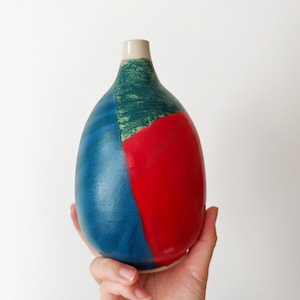May include: A hand-painted ceramic vase with a unique design. The vase features a color-blocked pattern with sections of blue, red, and green. The vase has a narrow neck and a rounded body. The vase is held in a person's hand.