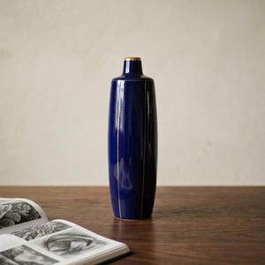May include: A tall, navy blue vase with gold accents. The vase has a narrow neck and a cylindrical body. Vertical gold lines run down the sides. The vase is on a wooden surface next to an open book with black and white illustrations.