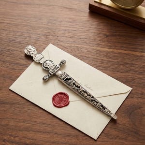 May include: A silver letter opener with an ornate handle rests on a cream-colored envelope sealed with a red wax stamp. The letter opener features detailed engravings and a series of sculpted faces. A small brass clock sits in the background on a wooden surface.