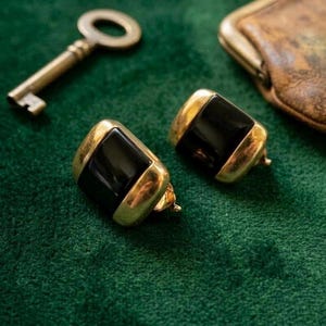 May include: A pair of gold-toned earrings with a rectangular black center stone. The earrings are set against a dark green velvet surface, with a vintage key and a small leather pouch in the background.