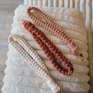 May include: Three macrame wrist lanyards in cream, terracotta, and peach colours, each with a rose gold swivel clasp. The lanyards are displayed on a soft, white, textured surface.