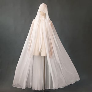 May include: A floor-length white hooded cloak made of sheer, lightweight fabric. The garment features a pointed hood and a simple tie closure at the neck, draping loosely from the shoulders to create a flowing, translucent silhouette.