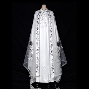 May include: White traditional Chinese Hanfu dress with a long, flowing skirt and a sheer overlay. The overlay and trim are adorned with black floral embroidery. The dress is displayed on a mannequin against a black background.