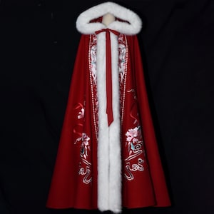 May include: A red hooded cloak with white faux fur trim and floral embroidery. The cloak features a hood, a front closure with a red ribbon, and decorative embroidery with pink and white flowers.