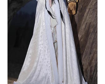 May include: A long, cream-colored cloak with a floral pattern and a faux fur trim. The cloak has a hood and is worn over a traditional outfit. The cloak is floor-length and appears to be made of a soft, flowing fabric.