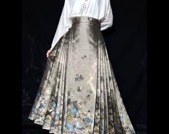 May include: An Asian-inspired outfit comprising a white top with embroidery and a long, pleated skirt. The skirt is a muted grey-green with a detailed floral and landscape pattern, accented with blue and gold. The outfit includes a wide, matching belt.