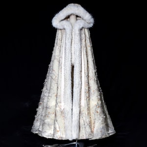 May include: A cream-coloured hooded cloak with a faux fur trim and hood. The cloak features a delicate floral and branch pattern. The interior and hood lining are also trimmed with faux fur. The cloak is displayed on a mannequin against a black background.