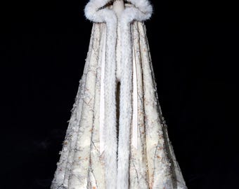 May include: A cream-colored hooded cloak with a faux fur trim and hood. The cloak features a delicate floral and branch pattern. The interior and hood lining are also trimmed with faux fur. The cloak is displayed on a mannequin against a black background.