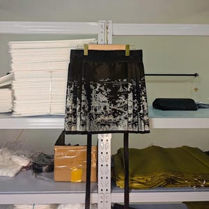 May include: Black pleated skirt with a white Asian-inspired landscape design. The skirt is hanging on a wooden hanger. The skirt has long black ties. The skirt is made of a shiny material.