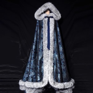 Könnte beinhalten: Ein Kapuzenmantel mit marineblauem Blumenmuster und grauem Kunstfellbesatz. Am Hals ist ein marineblaues Band gebunden. Der Umhang ist bodenlang und wirkt luxuriös.