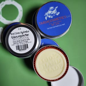 May include: Several small, round, silver tins with blue and white labels are arranged on a green surface. One tin is open, revealing a light yellow bar with "100% HAND MADE" embossed on it. The labels read "TARIACOSMETICS LLC" and "End Zone Hydrator Kokum Jojoba Bar".