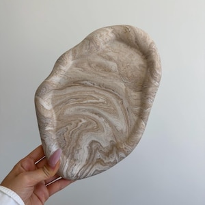 May include: A decorative tray with a unique, organic shape. The tray features a marbled design in shades of beige and white, creating a swirling pattern. The tray is held by a person, showcasing its size and design.