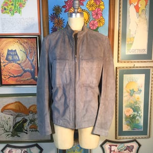 May include: A light gray suede jacket with a stand-up collar and a full-length zipper. The jacket has two chest pockets and is displayed on a mannequin. The jacket is a neutral color and could be worn by anyone.