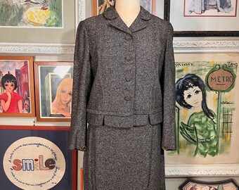 May include: A gray tweed skirt suit with a cropped jacket and a knee-length skirt. The jacket has a collar and three buttons. The skirt has a waistband and a straight silhouette.