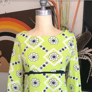 May include: A green dress with a white and navy blue geometric pattern. The dress has a boat neck and long sleeves. It has a black belt around the waist.