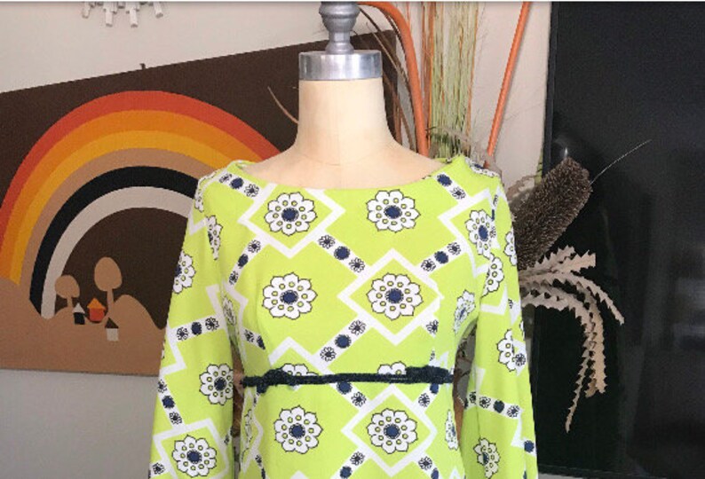 May include: A green dress with a white and navy blue geometric pattern. The dress has a boat neck and long sleeves. It has a black belt around the waist.