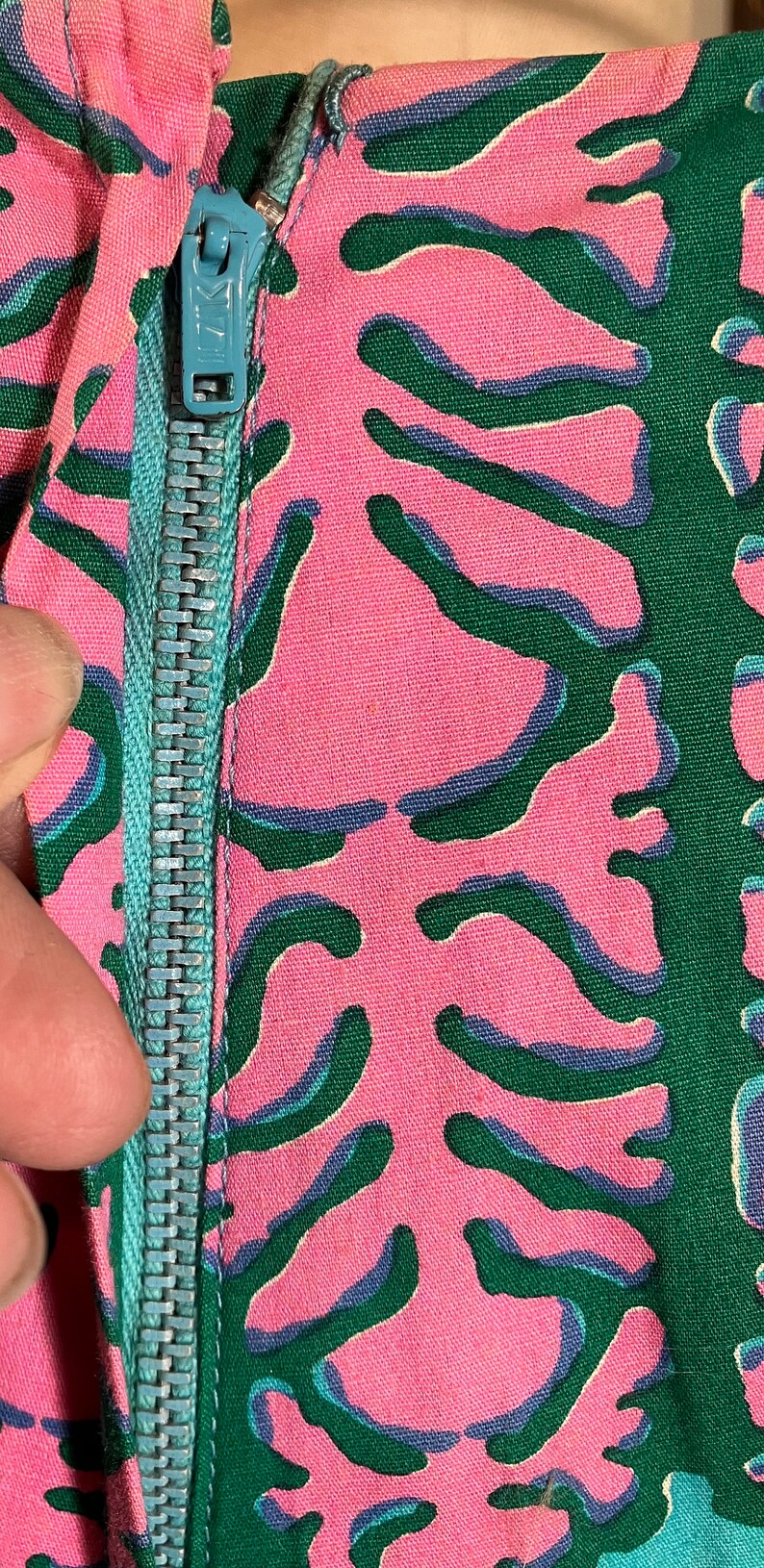 K&ouml;nnte beinhalten: Nahaufnahme eines t&uuml;rkisfarbenen Rei&szlig;verschlusses auf einem pink- und gr&uuml;n gemusterten Stoff.