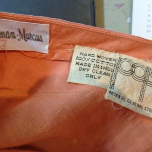 May include: A peach-colored garment with two white labels. The larger label reads "Neiman Marcus". The smaller label reads "Hand Woven 100% Cotton Made in India Dry Clean Only" and "Peter De General Stores".