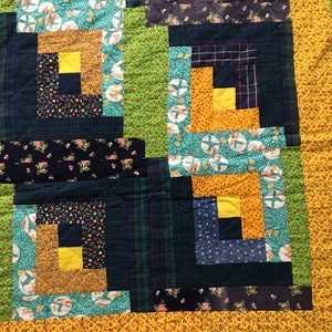 May include: A quilt made of various fabrics in shades of blue, green, and yellow. The quilt is made in a log cabin pattern with squares of different fabrics sewn together.