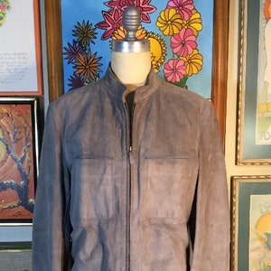 May include: A gray suede jacket with a zipper closure and two pockets on the front. The jacket is displayed on a mannequin in front of a wall with framed artwork.