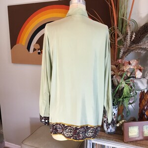 May include: A light green long-sleeved shirt with a brown and gold patterned trim around the bottom. The shirt is on a mannequin and is displayed against a brown background with a rainbow.