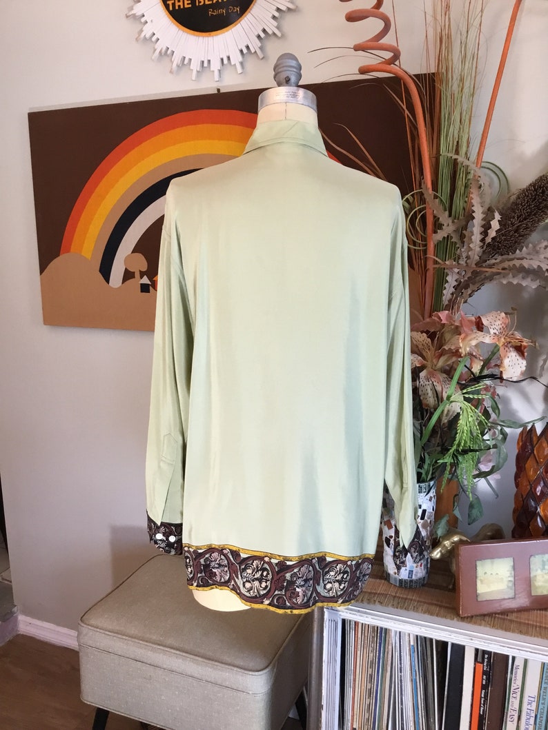 May include: A light green long-sleeved shirt with a brown and gold patterned trim around the bottom. The shirt is on a mannequin and is displayed against a brown background with a rainbow.